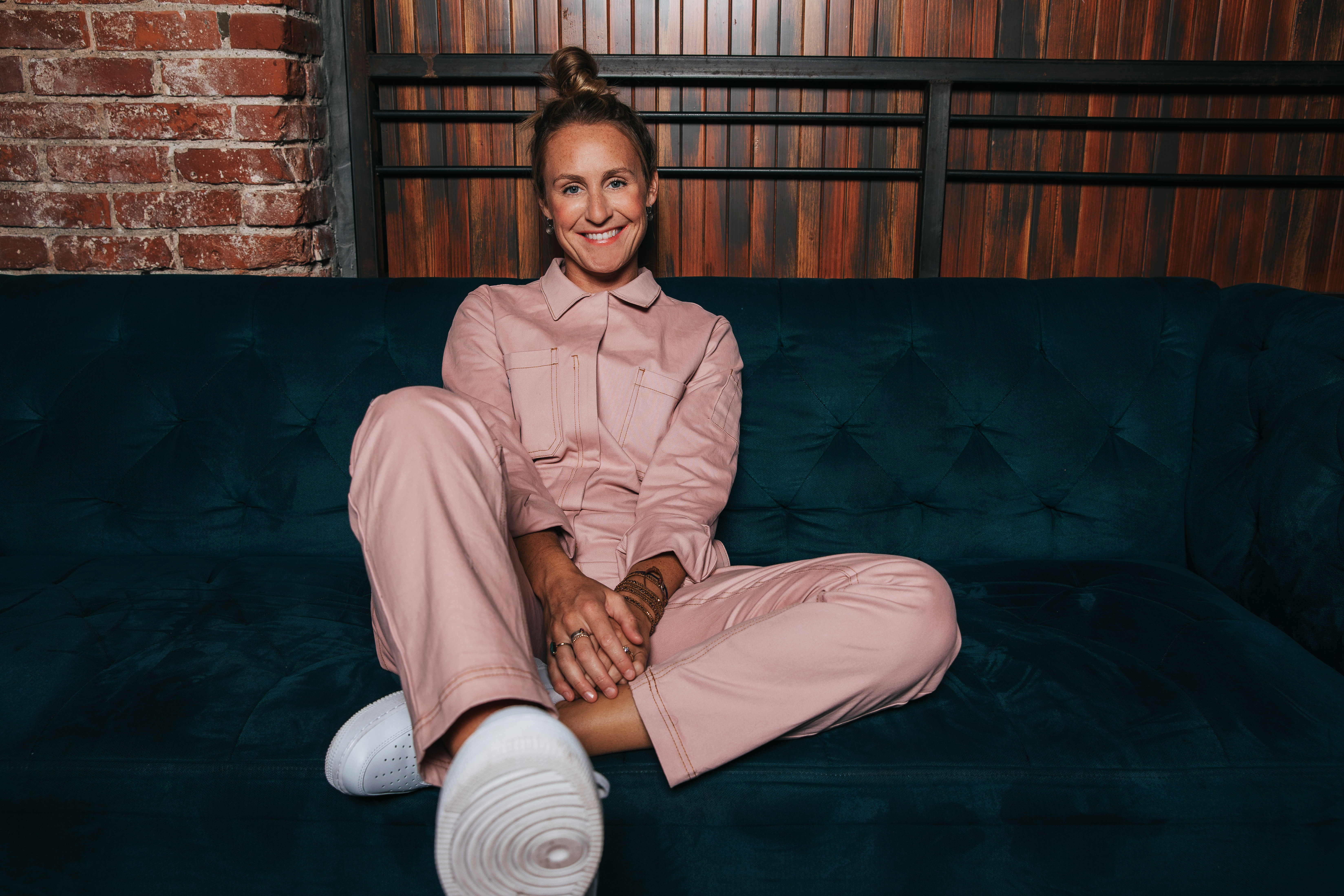 Person sitting cross-legged on a blue couch, smiling at the camera, wearing a pink jumpsuit and white sneakers, with a red brick and wooden slat background.