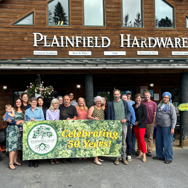 A large group of people pose in front of a building with "Plainfield Hardware Store" written on the top. The people hold a banner celebrating 50 years of being in business.