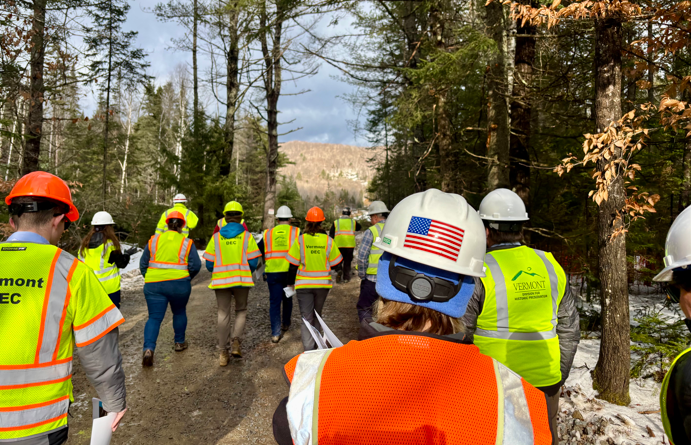 Several people in bright construction vests and hardhats walking in the woods.