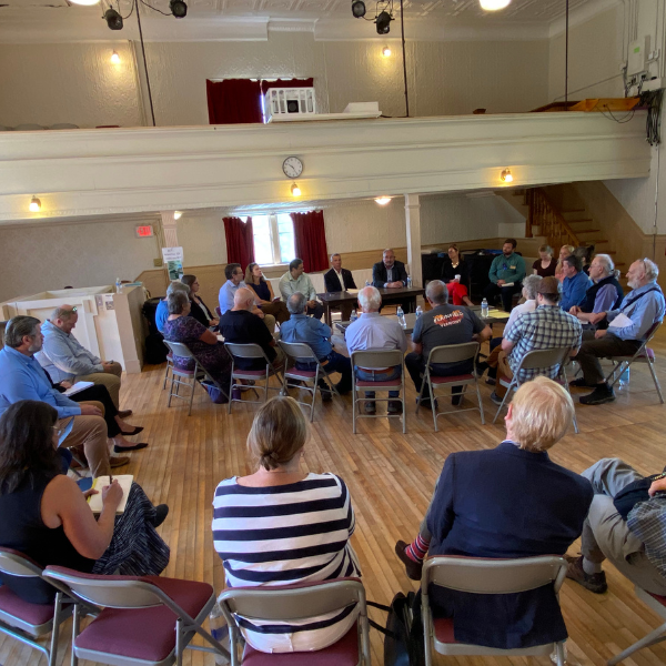 People are seated in chairs in a tight circle in a town hall. There are more people seated behind them in a second circle of chairs. 