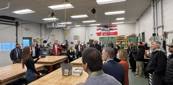 A group of people in a manufacturing shop floor, listening to one individual speak.