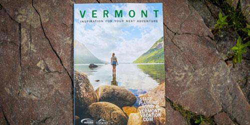 A photo of Vermont travel magazine cover featuring a person standing in a scenic lake surrounded by mountains lie on textured brick pavement