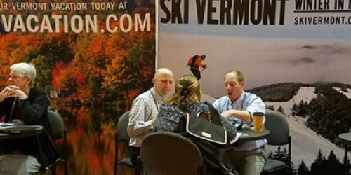 People engage in discussions at a travel booth promoting Vermont vacations, with a scenic backdrop and informational signs.